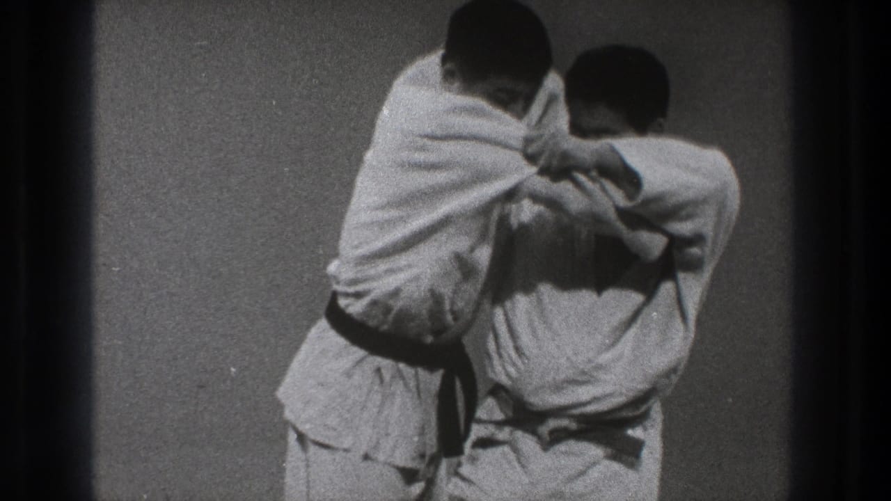 Two adult men in white gi continue a close-contact karate demonstration on a mat in the supplied 1971 Tokyo home-movie footage.
