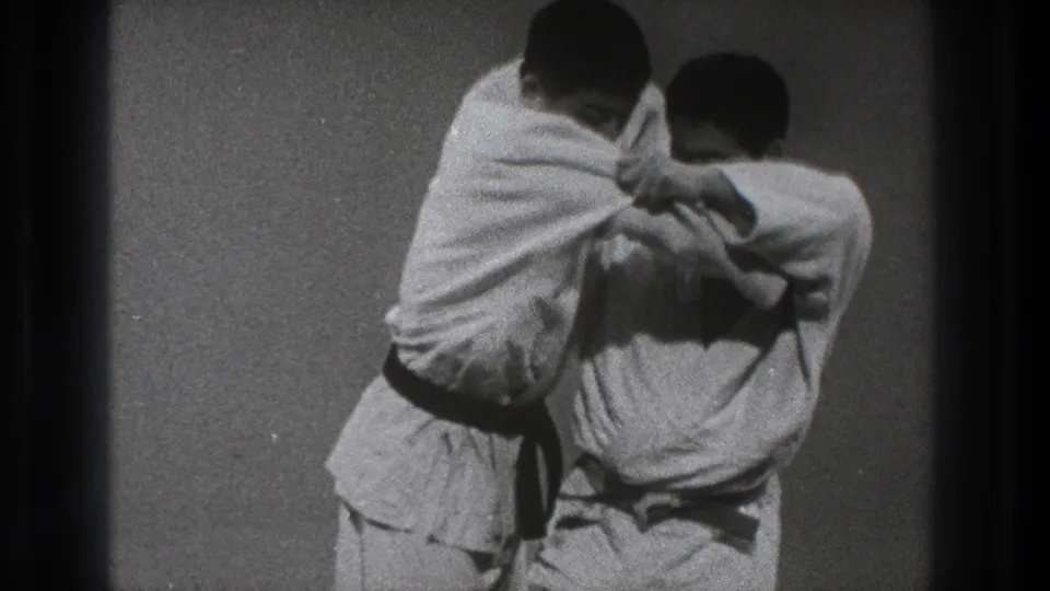 Two adult men in white gi continue a close-contact karate demonstration on a mat in the supplied 1971 Tokyo home-movie footage.