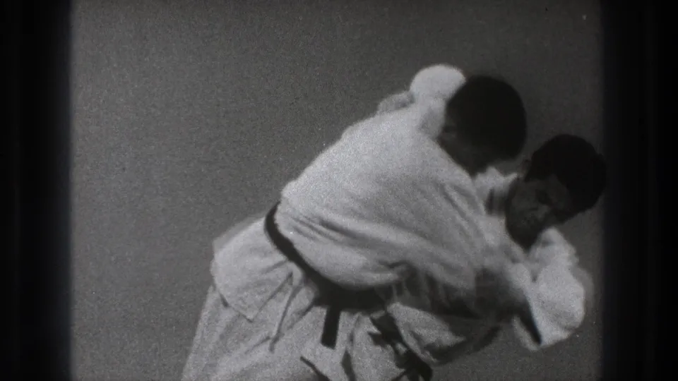 In Tokyo 1971 footage, two adult men in white gi and black belts practice a close judo-style hold on a padded mat.