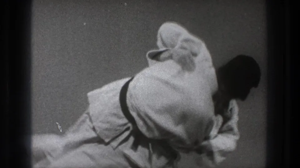 The black-and-white 8mm frame shows two adult men in gi low on the mat during a continued judo-style technique.