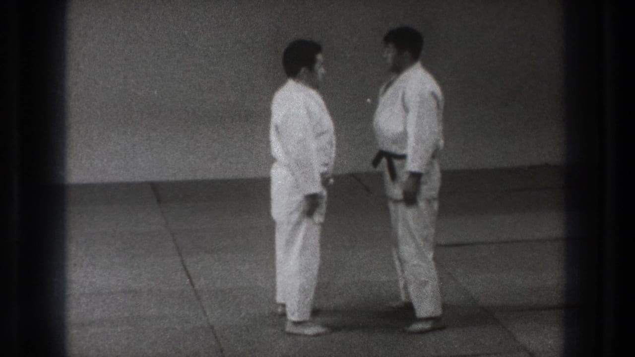 Two adult martial artists in white gi and black belts stand facing each other on a mat in Tokyo 1971 home-movie footage.