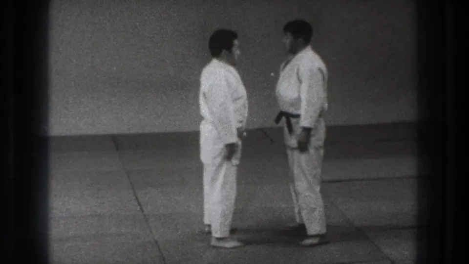 Two adult martial artists in white gi and black belts stand facing each other on a mat in Tokyo 1971 home-movie footage.