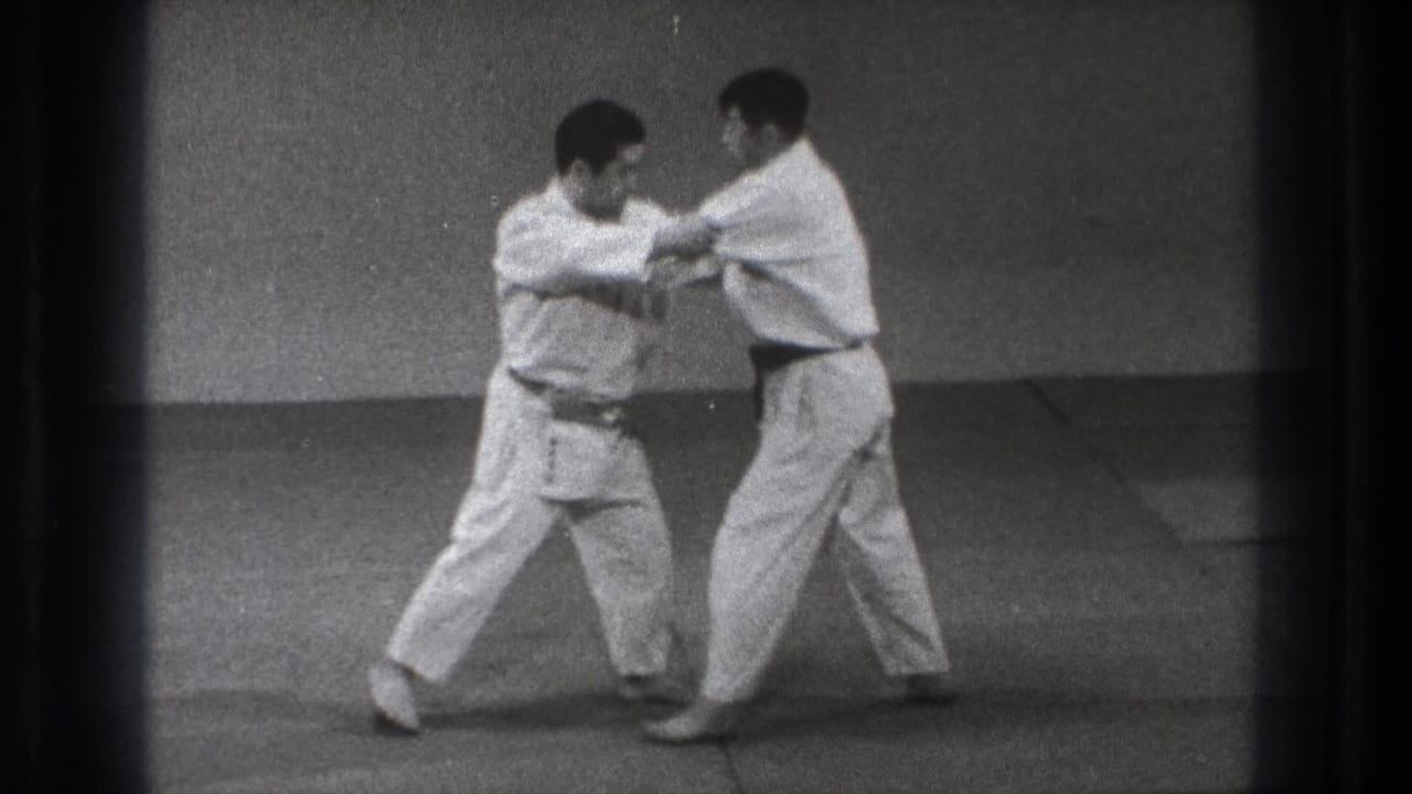 Two men in white gi practice a close arm parry or block on an indoor mat in the 1971 Tokyo footage.