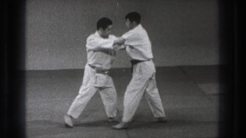 Two men in white gi practice a close arm parry or block on an indoor mat in the 1971 Tokyo footage.