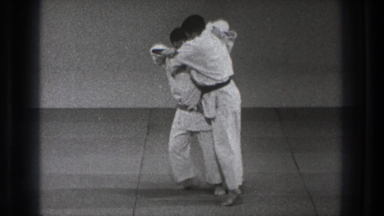In 1971 Tokyo home-movie footage, two judo practitioners in white gi and dark belts face each other at close range on a simple mat.