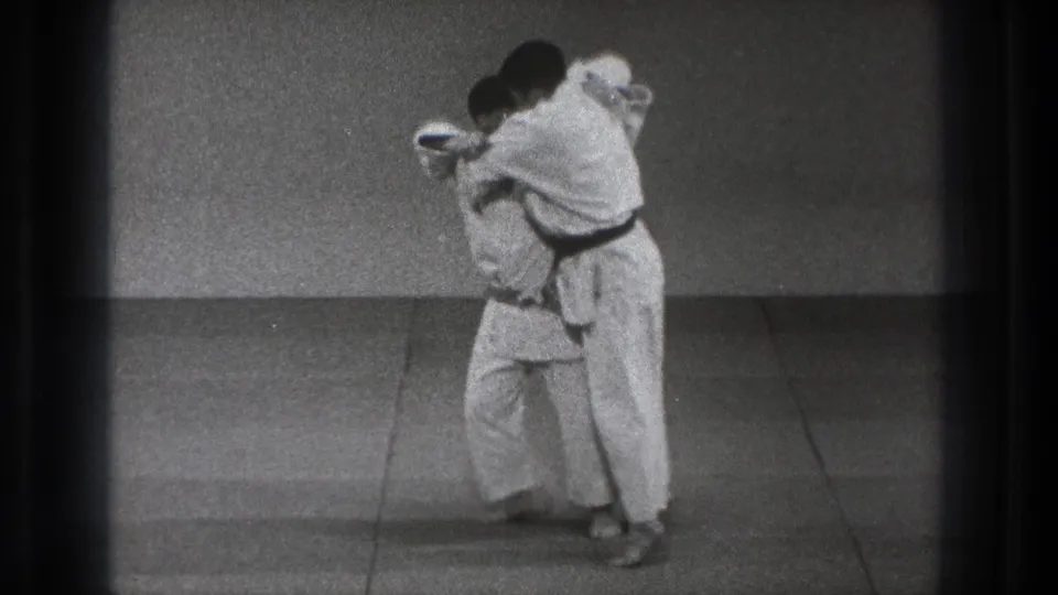 In 1971 Tokyo home-movie footage, two judo practitioners in white gi and dark belts face each other at close range on a simple mat.