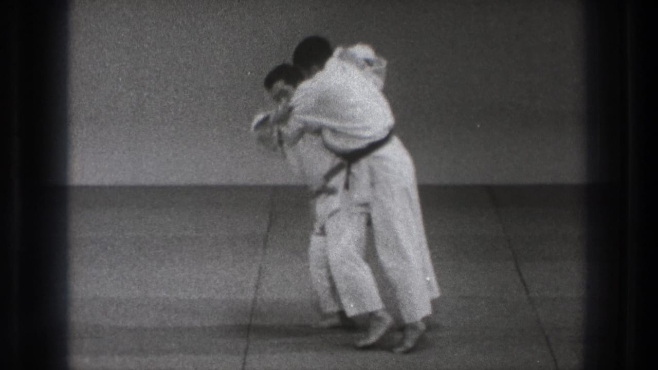 The paired judo demonstration continues as one practitioner shifts weight and begins to turn into the throw during the 1971 Tokyo clip.
