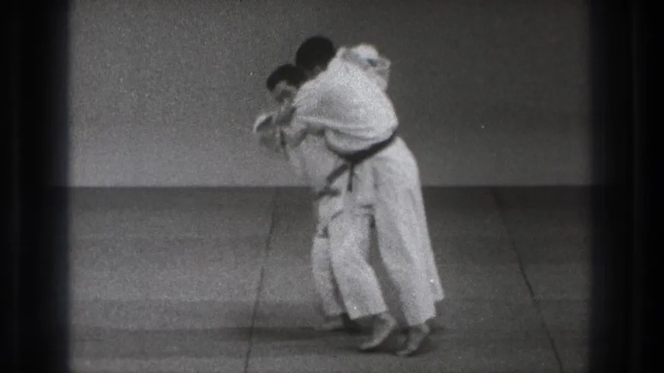 The paired judo demonstration continues as one practitioner shifts weight and begins to turn into the throw during the 1971 Tokyo clip.