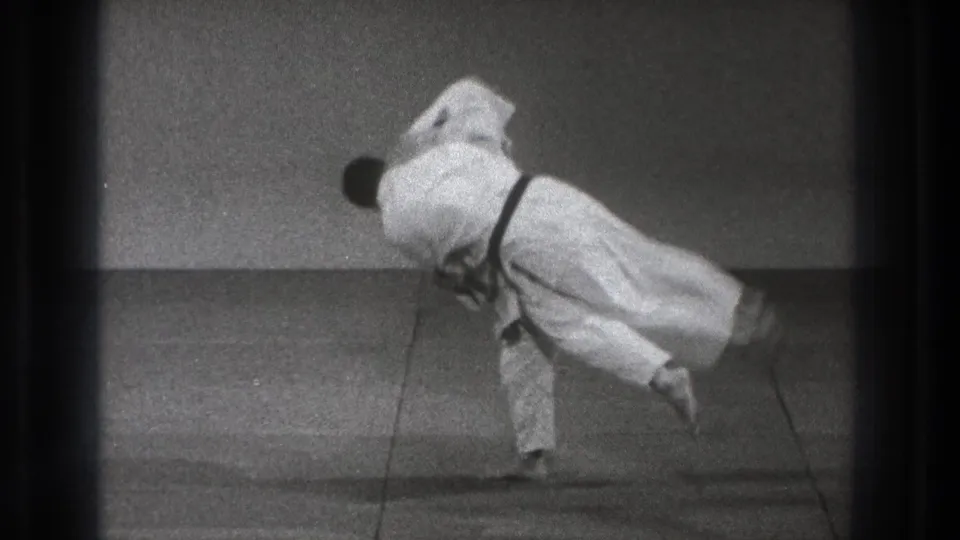 One practitioner is carried sideways through the throw, with both white gi blurred by the fast motion in the black-and-white 8mm frame.