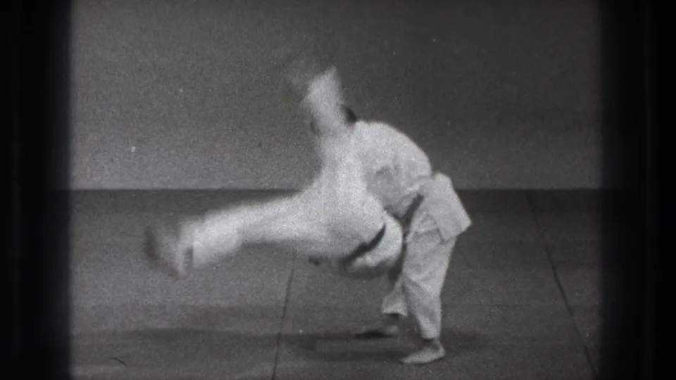 In Tokyo 1971 8mm footage, two adult men in white gi demonstrate a high martial arts kick on padded mats.