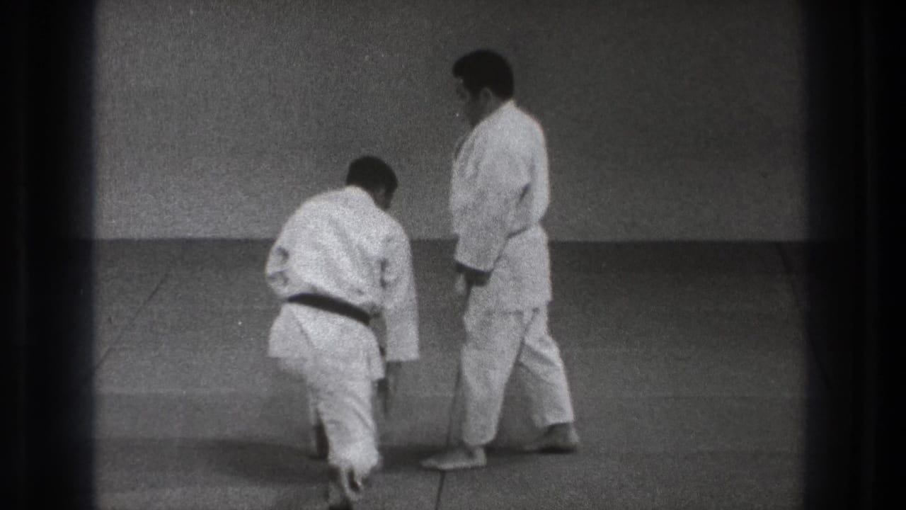 Two men in white judogi on a mat, one kneeling while the other stands nearby.