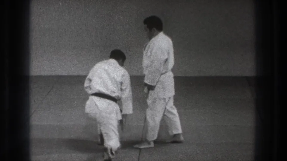 In Tokyo 1971 home-movie footage, two adult men in white judogi prepare on a mat as one kneels and the other stands.