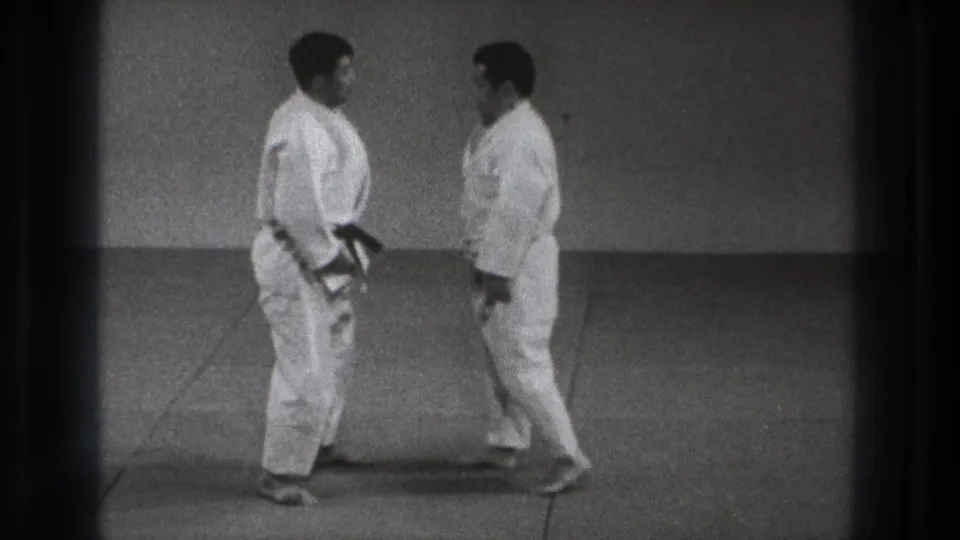 Two adult judo practitioners in white uniforms face one another on a mat in the 1971 Tokyo footage.
