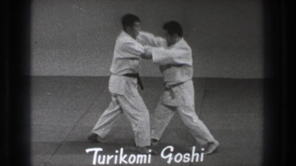 In 1971 Tokyo footage, two adult martial artists in white gi begin a paired Turikomi Goshi demonstration on an indoor mat.