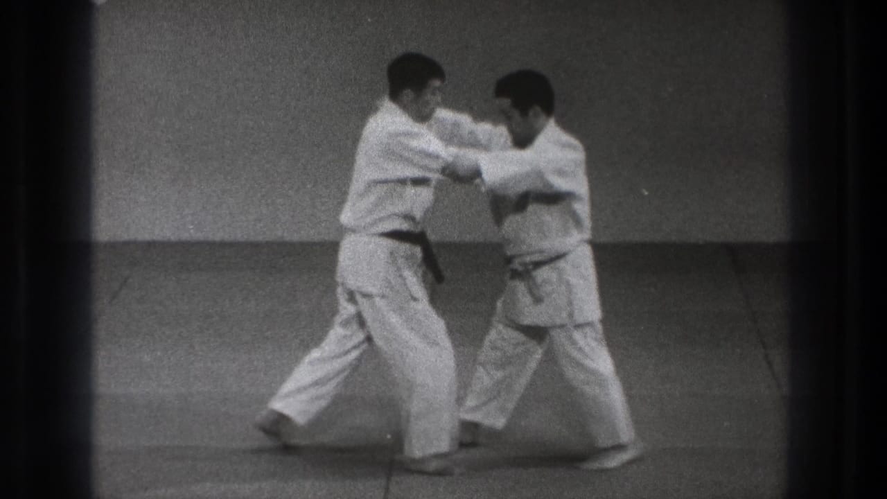 Two adult practitioners in white gi continue the 1971 Tokyo martial-arts demonstration, adjusting grips and foot placement on the mat.