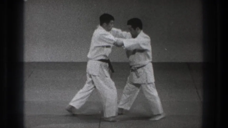 Two adult practitioners in white gi continue the 1971 Tokyo martial-arts demonstration, adjusting grips and foot placement on the mat.