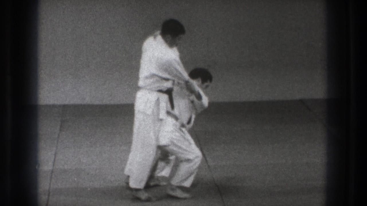 Two men in white gi begin a martial arts throw on a plain mat.