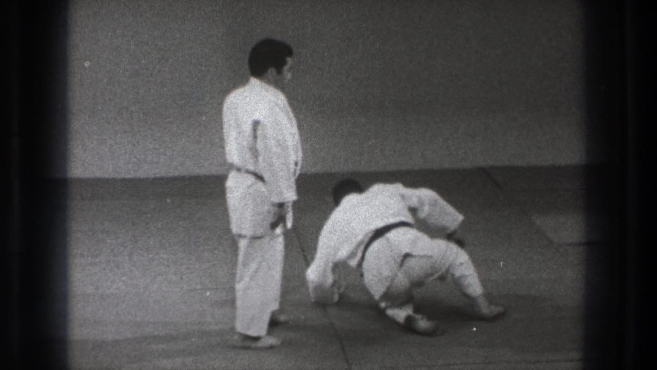 A karate practitioner stands nearby as his partner rolls and begins to recover on the indoor mat in 1971 Tokyo footage.