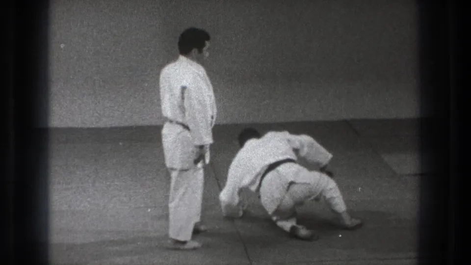 A karate practitioner stands nearby as his partner rolls and begins to recover on the indoor mat in 1971 Tokyo footage.