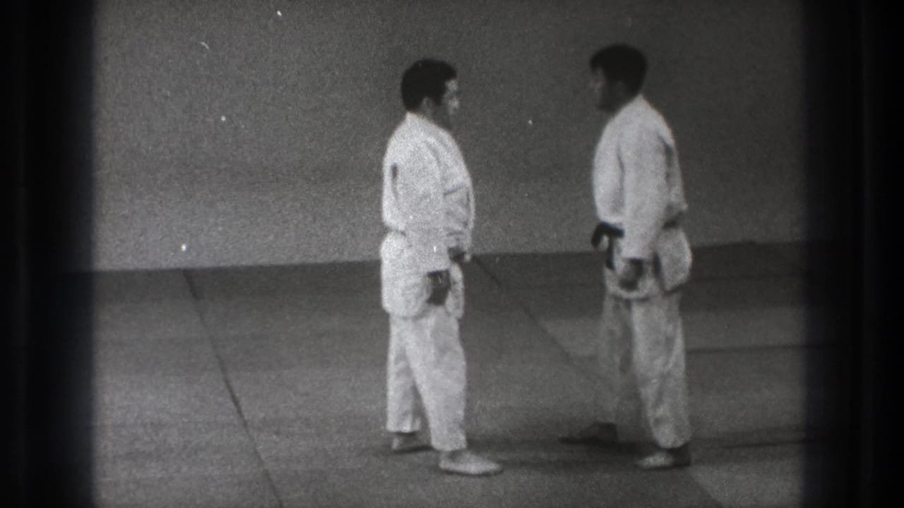 Two men in white judo uniforms stand face to face in a Tokyo dojo, one wearing a dark belt.
