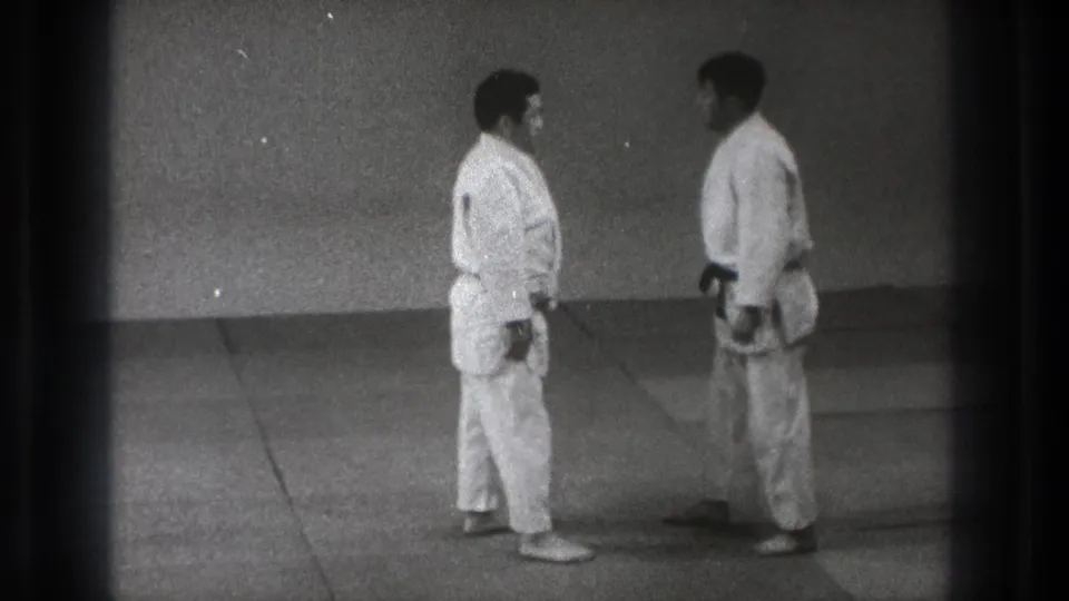 Two men in white judo uniforms stand face to face in a Tokyo dojo, one wearing a dark belt.
