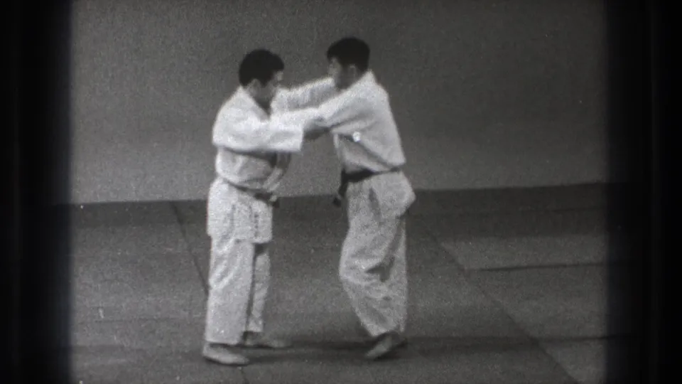 In metadata-supported 1971 Tokyo, two adult men in white uniforms and dark belts begin a grappling practice exchange on a mat.
