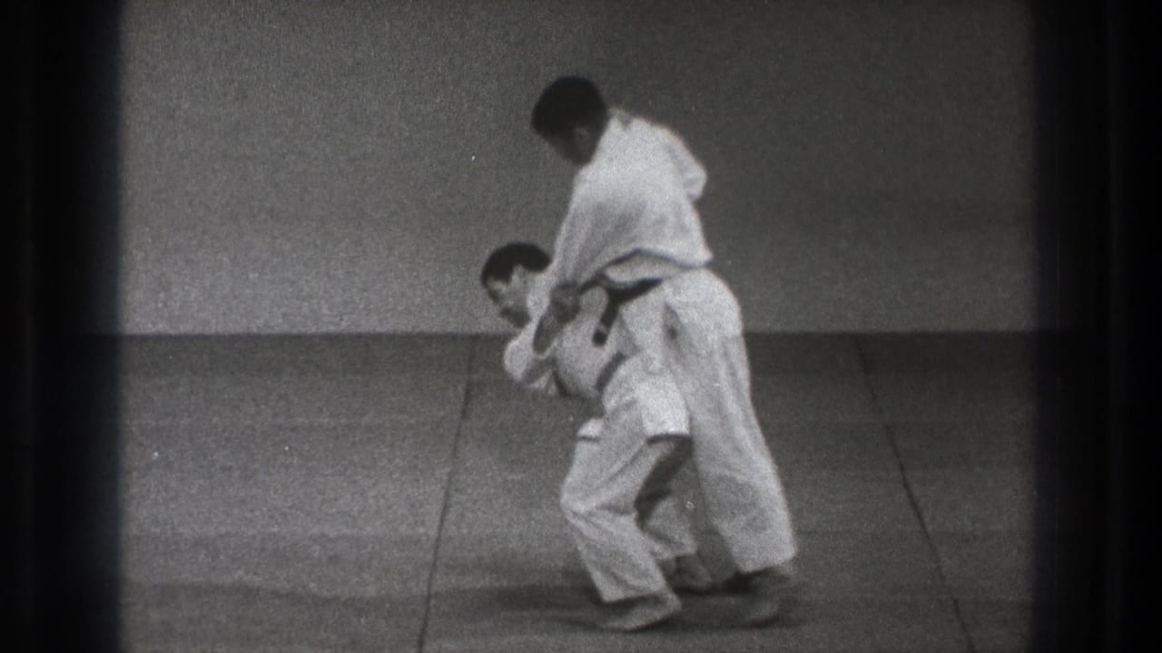 A man in a white martial-arts uniform bends forward while his partner stands over the hold near the end of the 1971 Tokyo sequence.