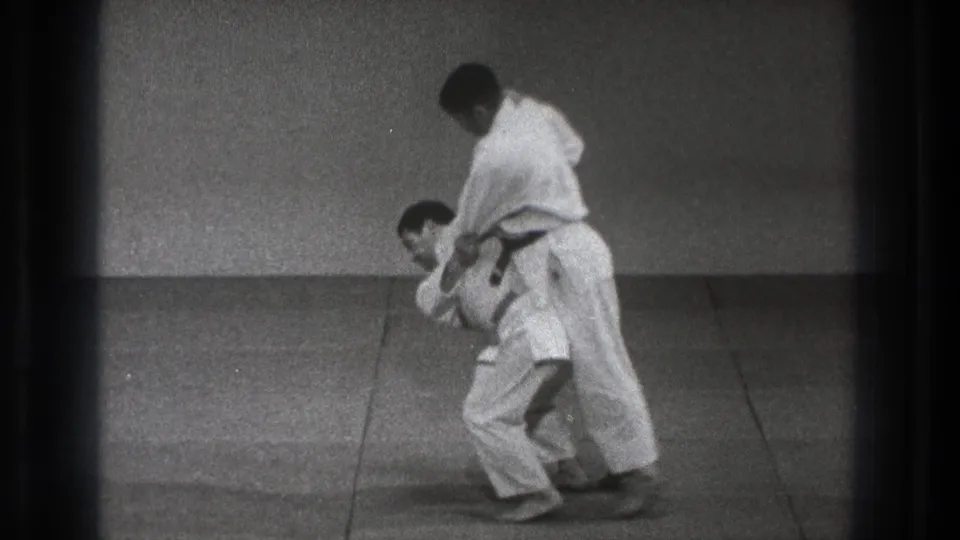 A man in a white martial-arts uniform bends forward while his partner stands over the hold near the end of the 1971 Tokyo sequence.