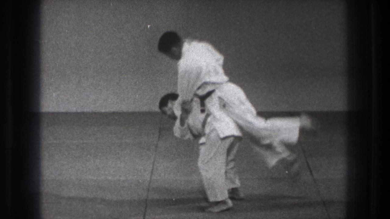 Two judo practitioners in white uniforms begin a throw on a grey mat.