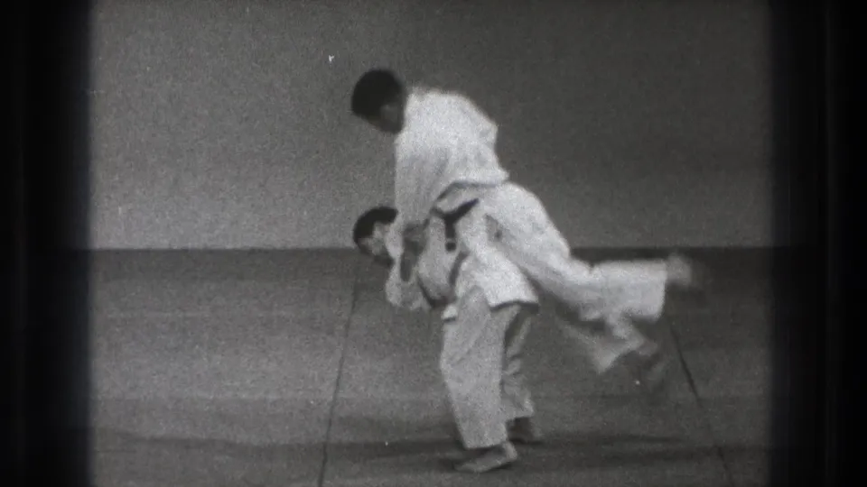 In 1971 Tokyo 8mm footage, two men in white judogi start a judo throw on a plain grey mat.