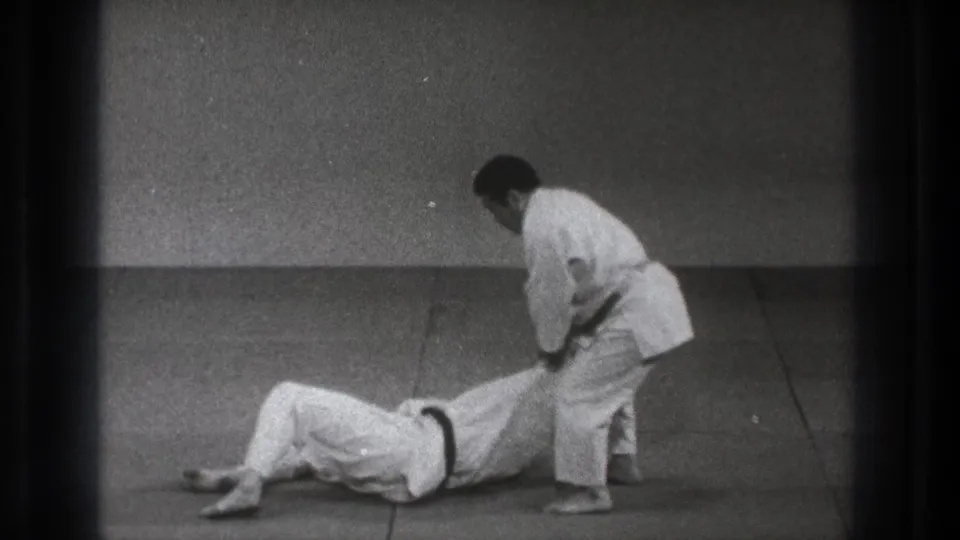 After the throw, one man lies on the grey mat as the standing practitioner keeps hold of his arm in the Tokyo dojo scene.