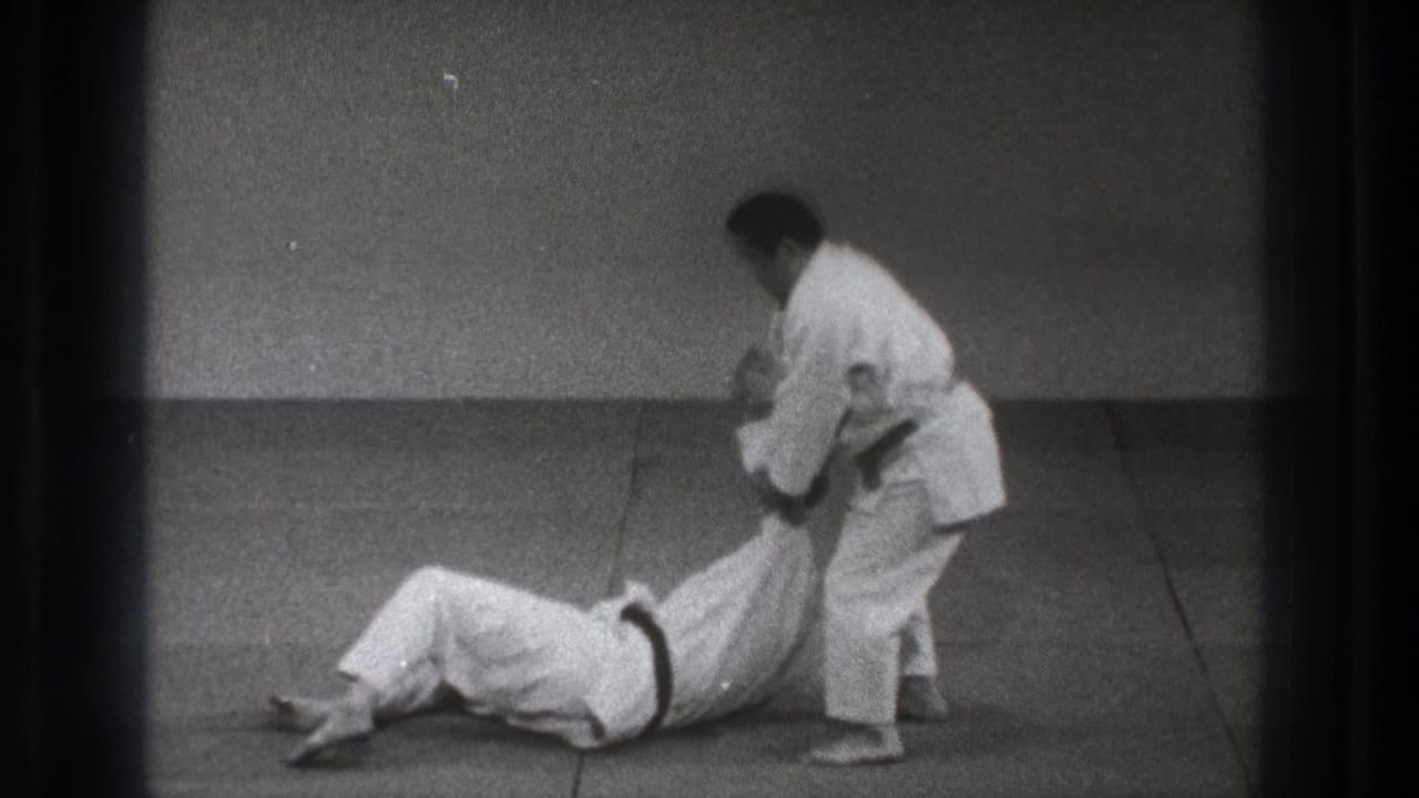 In 1971 Tokyo judo practice footage, one adult man in a white uniform pulls another seated opponent by the arm on a tatami mat.