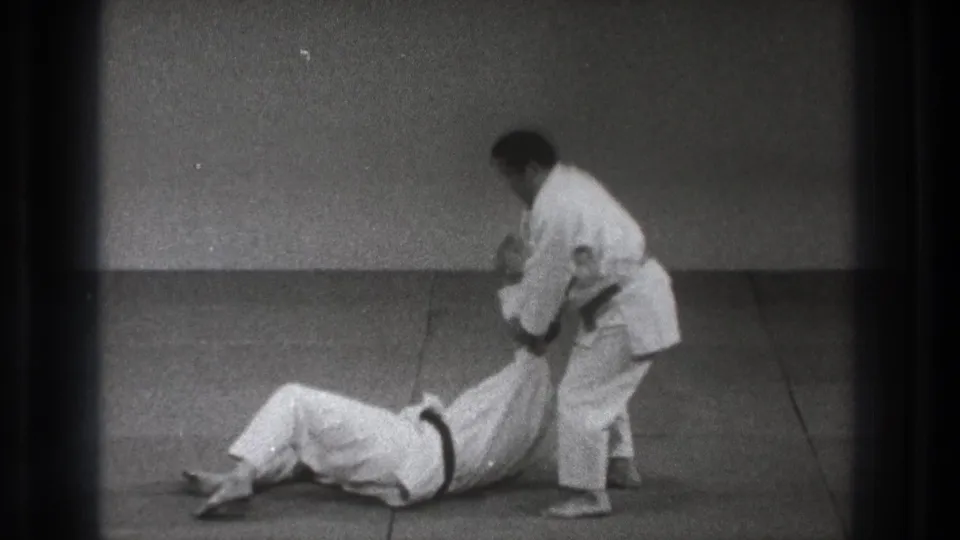 In 1971 Tokyo judo practice footage, one adult man in a white uniform pulls another seated opponent by the arm on a tatami mat.
