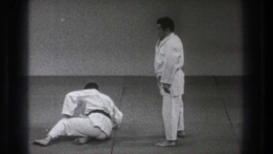During a 1971 Tokyo dojo practice moment, one adult judoka lies or sits low on the mat while the other stands watching nearby.