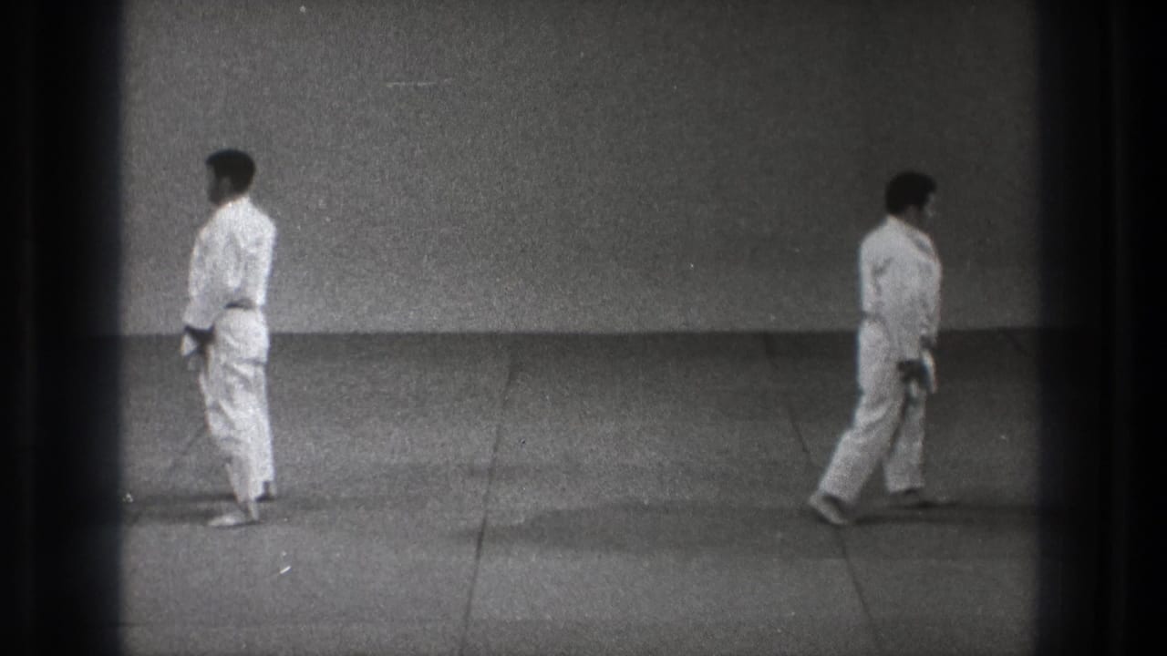 Man in a white gi stands on a plain mat within dark 8mm film borders.