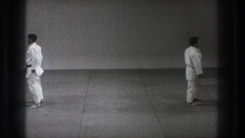 Two men in white gi appear across a simple mat in the 1971 Tokyo martial arts footage, framed by dark 8mm borders.