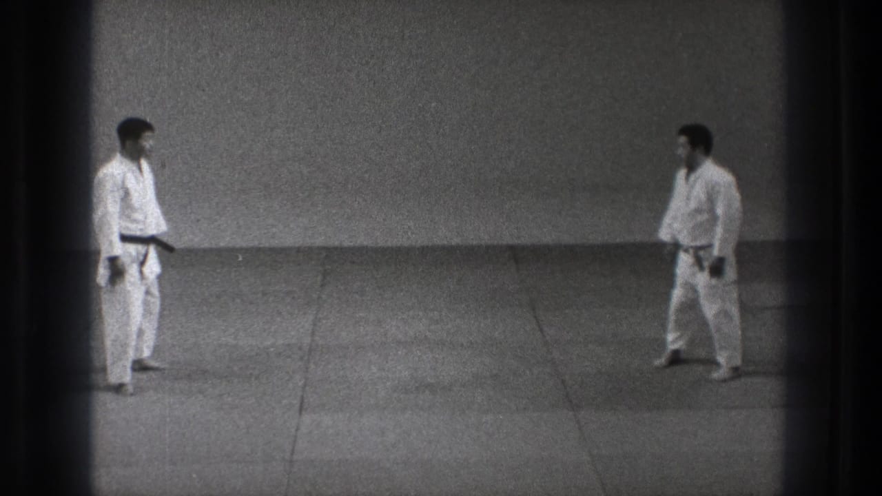 In a plain dojo setting, two judo practitioners in white uniforms face each other across the mat.