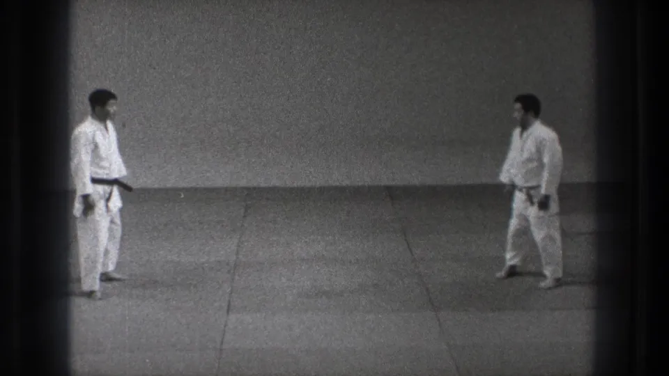 In a plain dojo setting, two judo practitioners in white uniforms face each other across the mat.