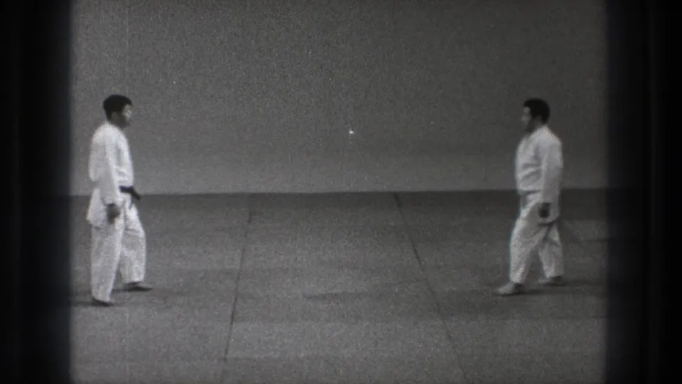 Two judo practitioners in white judogi walk toward each other in a black-and-white 8mm frame.