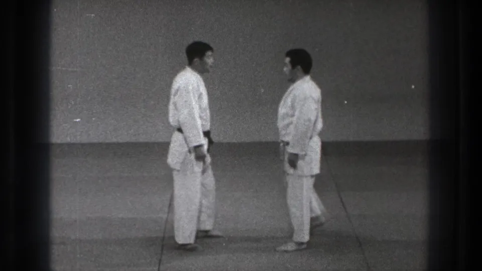 In 1971 Tokyo footage, two adult men in white gi stand facing each other on tatami-style mats before beginning a martial arts technique.