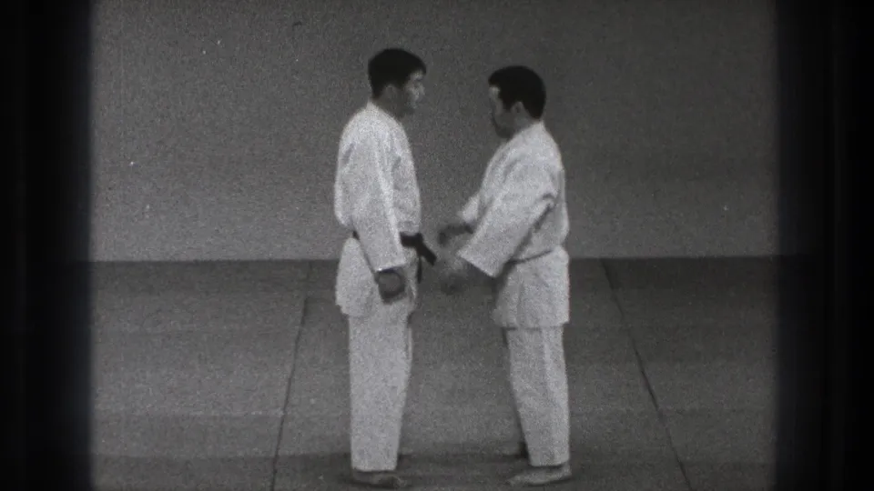 In the Tokyo dojo scene, two adult practitioners stand close as a controlled hand or grip movement begins during martial arts practice.
