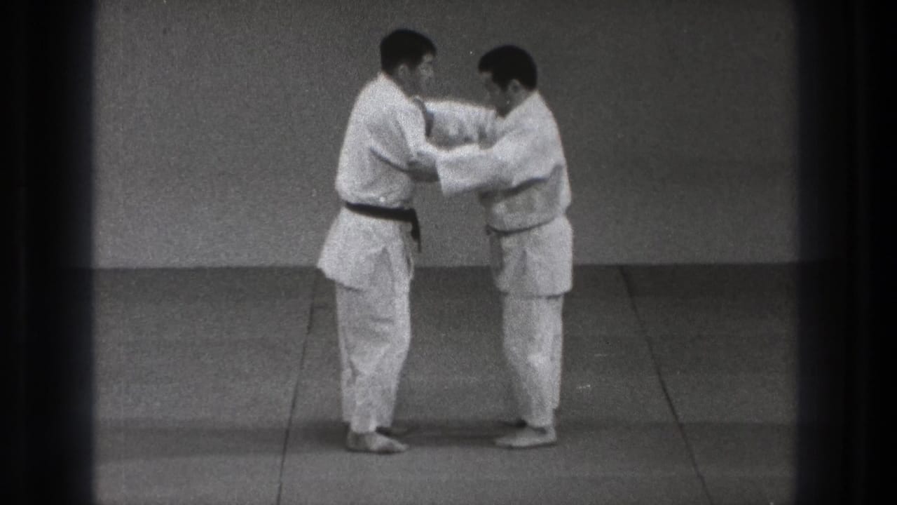 Two adult men in white gi hold arm positions on a tatami-style mat, demonstrating a controlled martial arts contact technique in Tokyo.