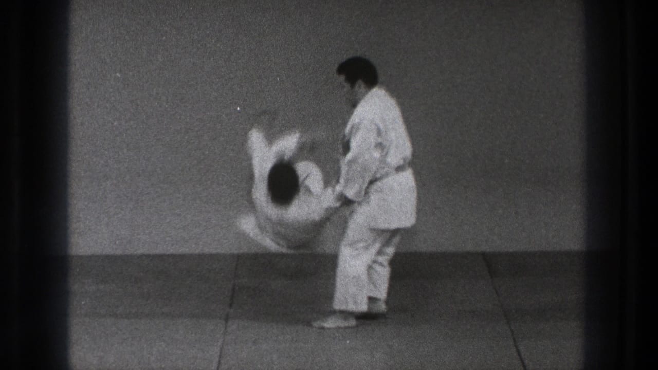 Two men in white gi practice a judo throw on floor mats.
