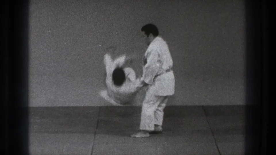 In Tokyo 1971, two adult martial artists in white gi are captured mid-throw on a plain dojo mat.