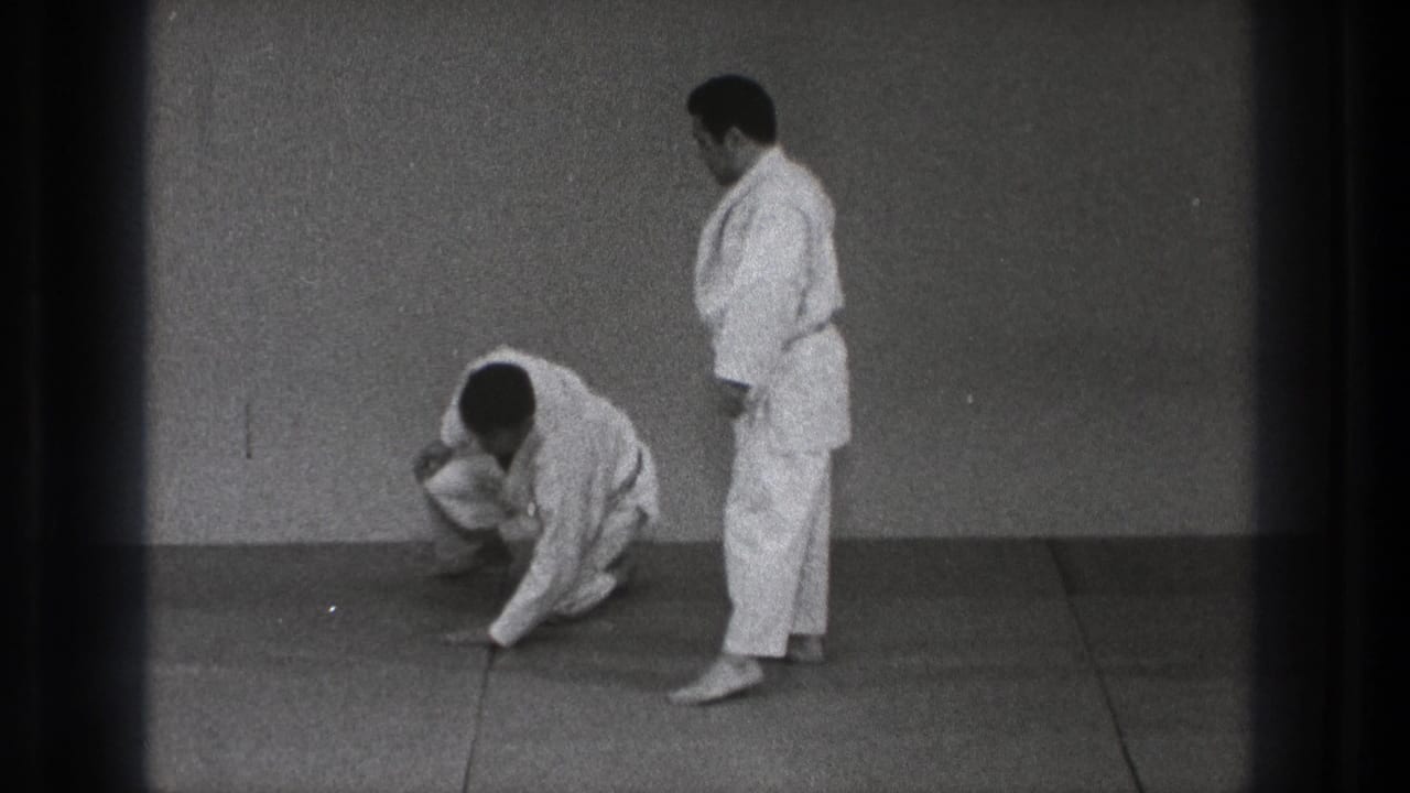 One adult martial artist kneels or rises from the mat as his partner stands nearby in the simple Tokyo dojo setting.