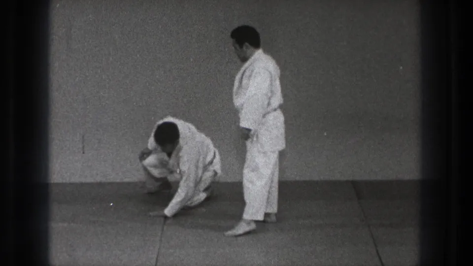 One adult martial artist kneels or rises from the mat as his partner stands nearby in the simple Tokyo dojo setting.