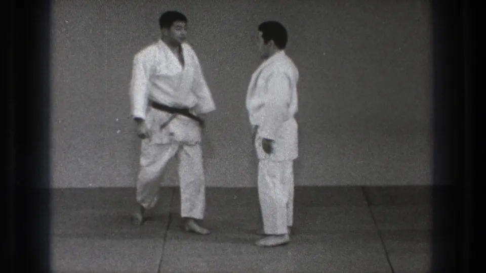 In 1971 Tokyo footage, two adult men in white judo gis face each other on a training mat, one wearing a black belt.