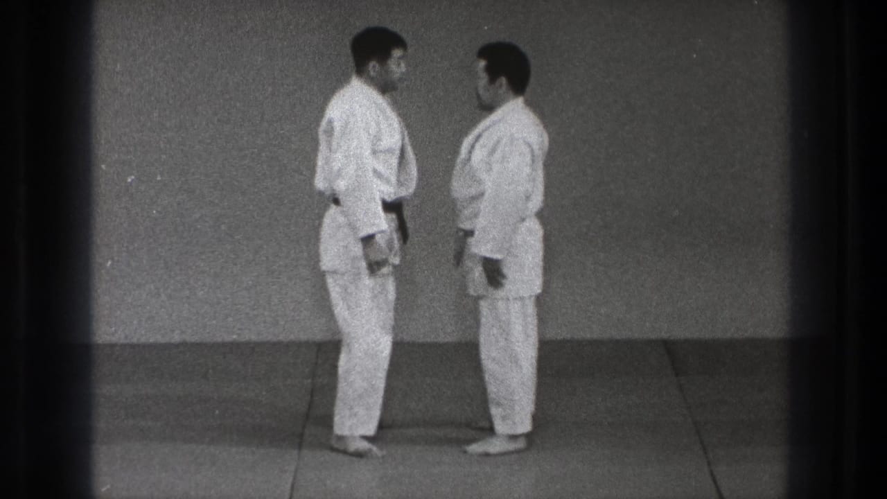 Two men in white judo uniforms stand close on a mat in the Tokyo training scene, facing one another in profile.