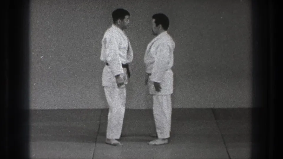 Two men in white judo uniforms stand close on a mat in the Tokyo training scene, facing one another in profile.