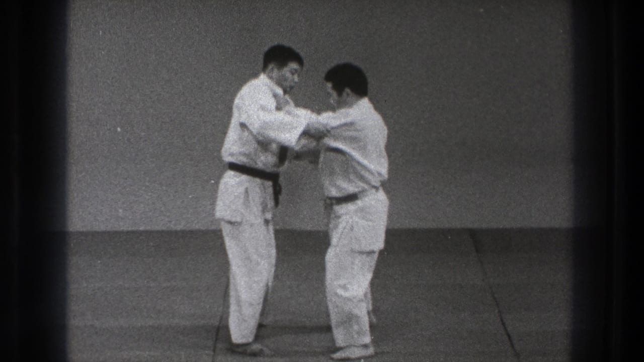 Two adult judo practitioners continue a close grappling technique on the mat, with one man wearing a black belt.